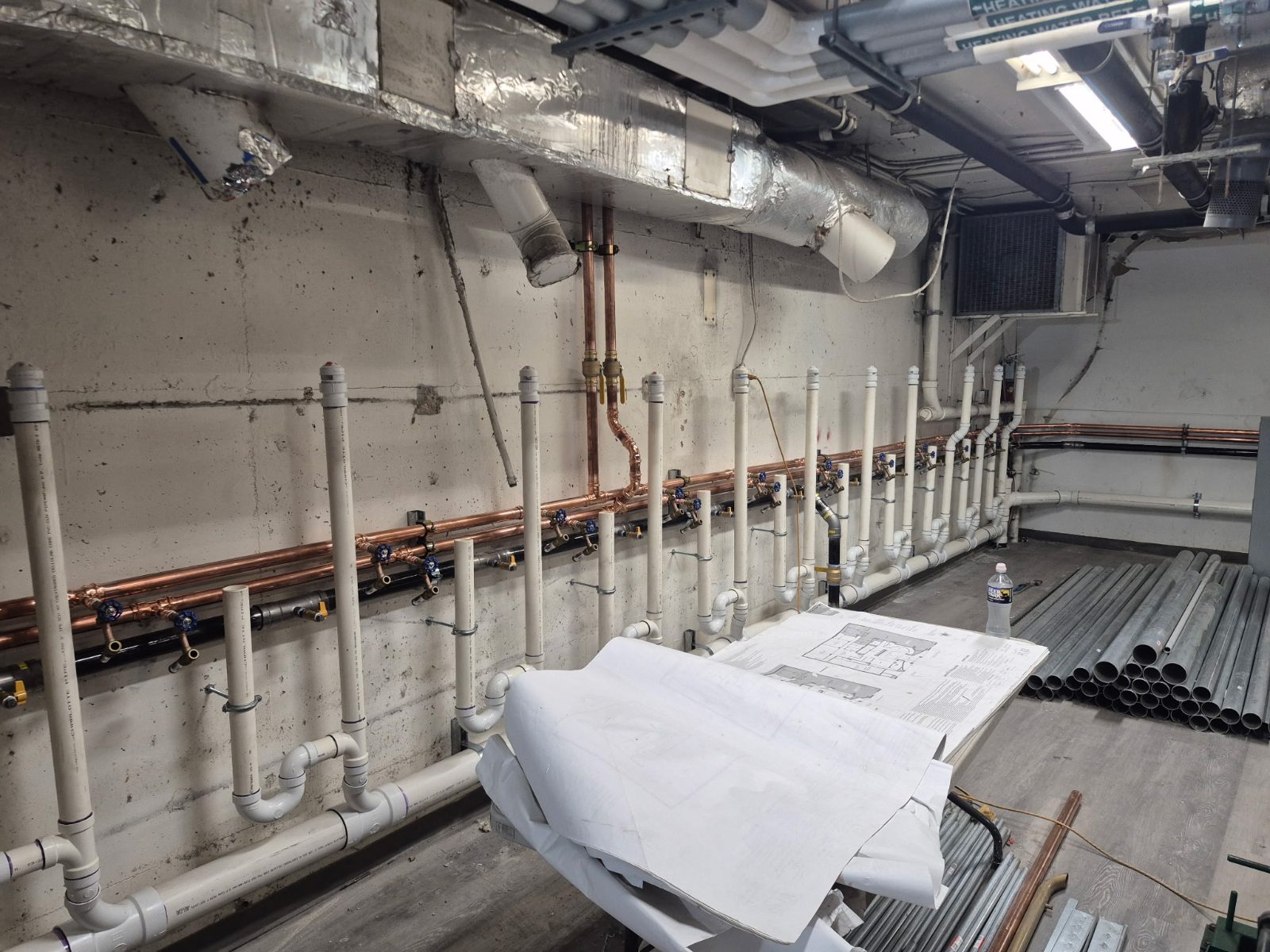 A mechanical room mid-build — copper mains, risers, duct overhead and the prints spread out on the bench